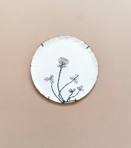 A round white enamel plate with a hand-drawn illustration of a clover plant in black.